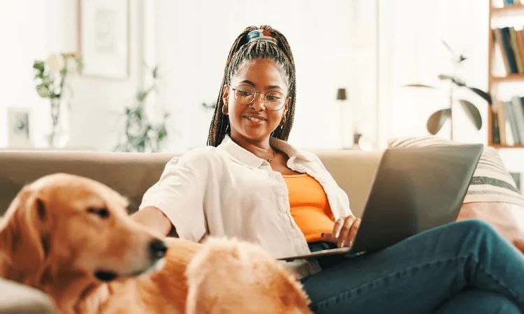 A female owner on the sofa with a laptop gently stroking her golden retrievers head