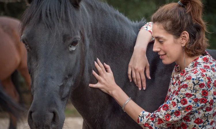A female rider in a floral shirt soothingly stoking a black horse's neck.